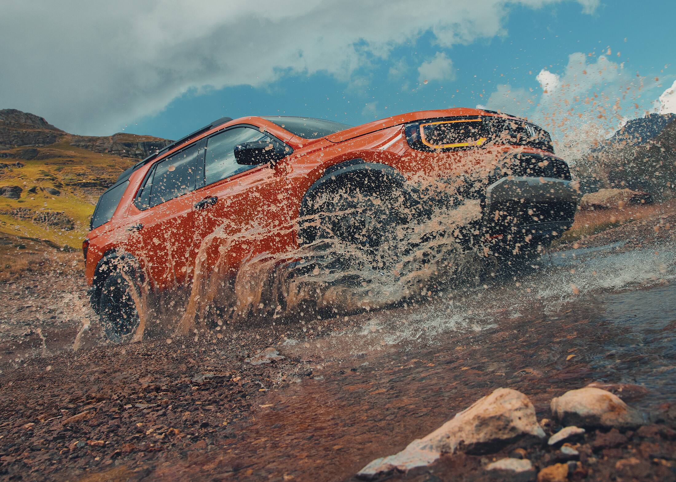 7/8 front passenger-side view of the 2026 Honda Passport TrailSport Elite in Sunset Orange, shown driving through a muddy water puddle with muddy water splashing up on the passenger side.